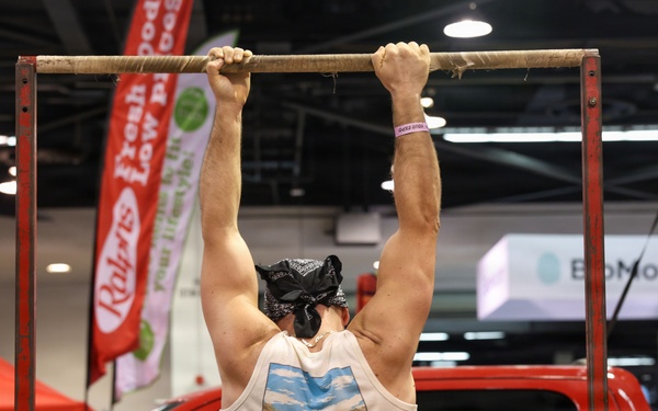 A muscular man wearing a Columbia tank top and black bandana is performing pull-ups on a makeshift bar at what appears to be an indoor expo or trade show. Two assistants are holding the metal frame steady on either side. The background features vendor booths, banners including 'Raley's' and 'BioMotion', and a red pickup truck on display.