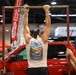 A muscular man wearing a Columbia tank top and black bandana is performing pull-ups on a makeshift bar at what appears to be an indoor expo or trade show. Two assistants are holding the metal frame steady on either side. The background features vendor booths, banners including 'Raley's' and 'BioMotion', and a red pickup truck on display.