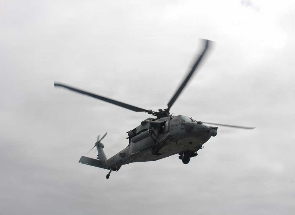 A military helicopter is captured in flight against an overcast gray sky. The aircraft appears to be a Sikorsky UH-60 Black Hawk or similar variant, painted in a gray military finish. The rotor blades show motion blur, indicating active flight.