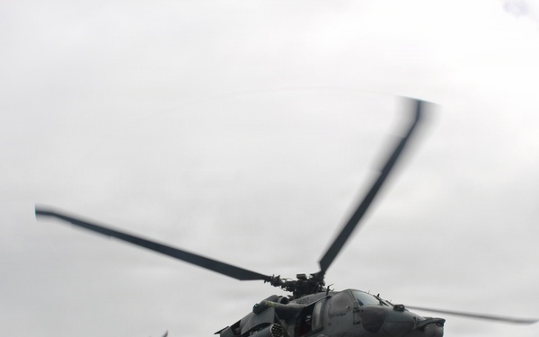 A military helicopter is captured in flight against an overcast gray sky. The aircraft appears to be a Sikorsky UH-60 Black Hawk or similar variant, painted in a gray military finish. The rotor blades show motion blur, indicating active flight.