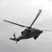 A military helicopter is captured in flight against an overcast gray sky. The aircraft appears to be a Sikorsky UH-60 Black Hawk or similar variant, painted in a gray military finish. The rotor blades show motion blur, indicating active flight.