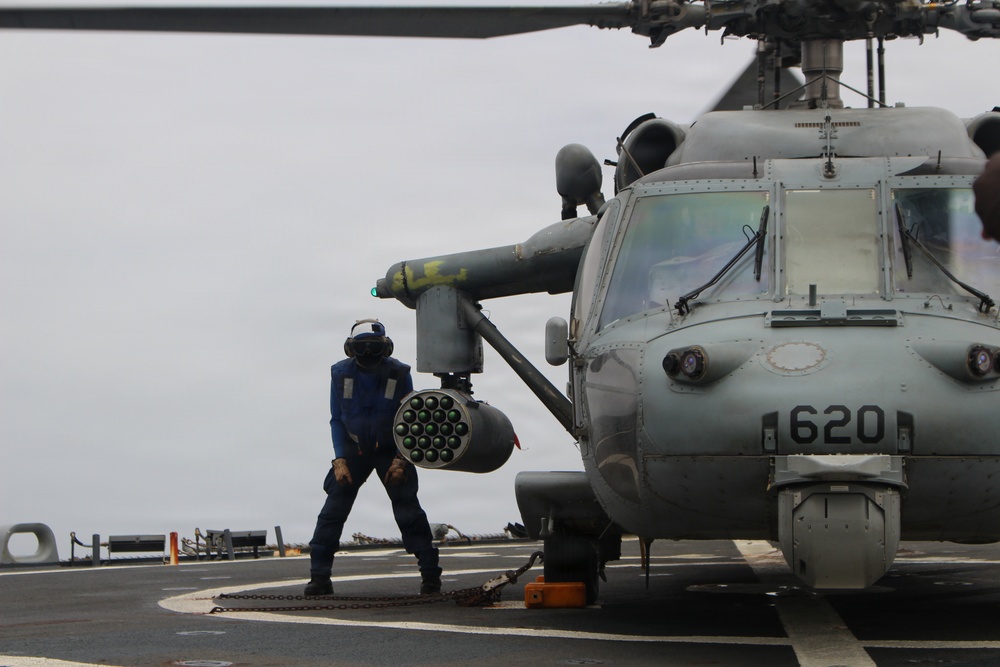 A naval deck crew member in blue coveralls and a cranial helmet is working alongside a military helicopter numbered '620' on what appears to be a ship's flight deck. The helicopter is equipped with a rocket pod launcher mounted on its stub wing. The scene takes place under overcast skies at sea.