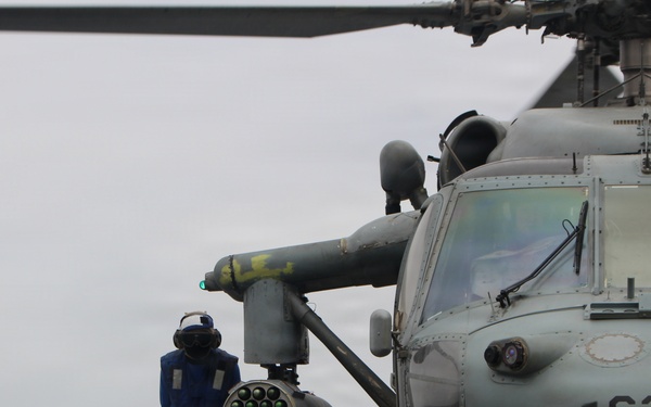 A naval deck crew member in blue coveralls and a cranial helmet is working alongside a military helicopter numbered '620' on what appears to be a ship's flight deck. The helicopter is equipped with a rocket pod launcher mounted on its stub wing. The scene takes place under overcast skies at sea.