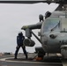 A naval deck crew member in blue coveralls and a cranial helmet is working alongside a military helicopter numbered '620' on what appears to be a ship's flight deck. The helicopter is equipped with a rocket pod launcher mounted on its stub wing. The scene takes place under overcast skies at sea.
