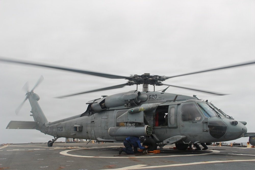A U.S. Navy helicopter, marked with the number '620', is positioned on a ship's flight deck with its rotors spinning. Ground crew personnel are visible beneath the aircraft, appearing to perform maintenance or pre-flight checks. The helicopter is armed with what appears to be an AGM-114 Hellfire missile system mounted on its side.