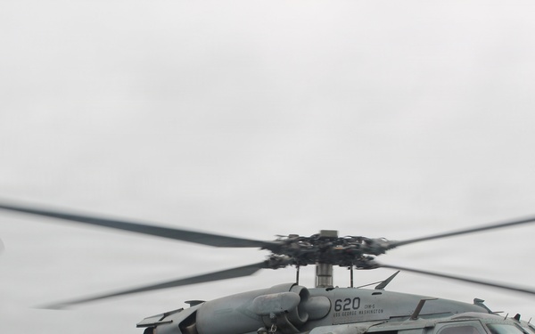 A U.S. Navy helicopter, marked with the number '620', is positioned on a ship's flight deck with its rotors spinning. Ground crew personnel are visible beneath the aircraft, appearing to perform maintenance or pre-flight checks. The helicopter is armed with what appears to be an AGM-114 Hellfire missile system mounted on its side.