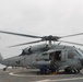 A U.S. Navy helicopter, marked with the number '620', is positioned on a ship's flight deck with its rotors spinning. Ground crew personnel are visible beneath the aircraft, appearing to perform maintenance or pre-flight checks. The helicopter is armed with what appears to be an AGM-114 Hellfire missile system mounted on its side.