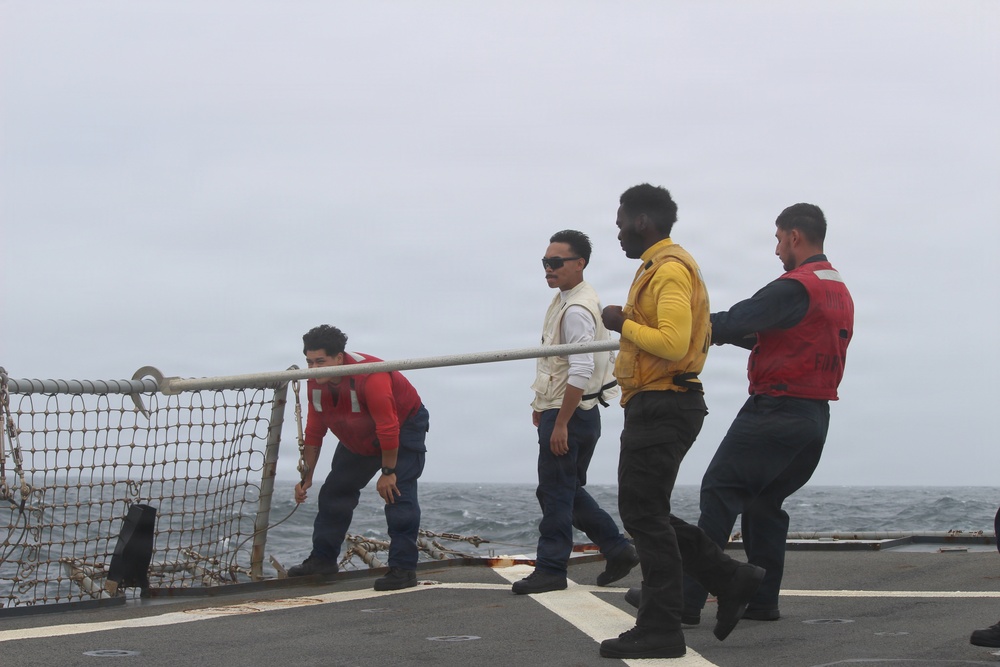 Four personnel are working together on the deck of a naval vessel at sea, handling a metal pole or bar in a coordinated effort. The crew members are wearing safety vests in red and yellow, suggesting an operational or deck maintenance activity. The overcast sky and choppy ocean waters are visible in the background.
