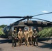 Five military personnel are posing in front of a dark olive/black UH-60 Black Hawk helicopter on a paved surface. The group includes individuals wearing U.S. Army multicam uniforms and what appear to be law enforcement or aviation flight suits. The setting appears to be an outdoor staging area with trees visible in the background on a clear, sunny day.
