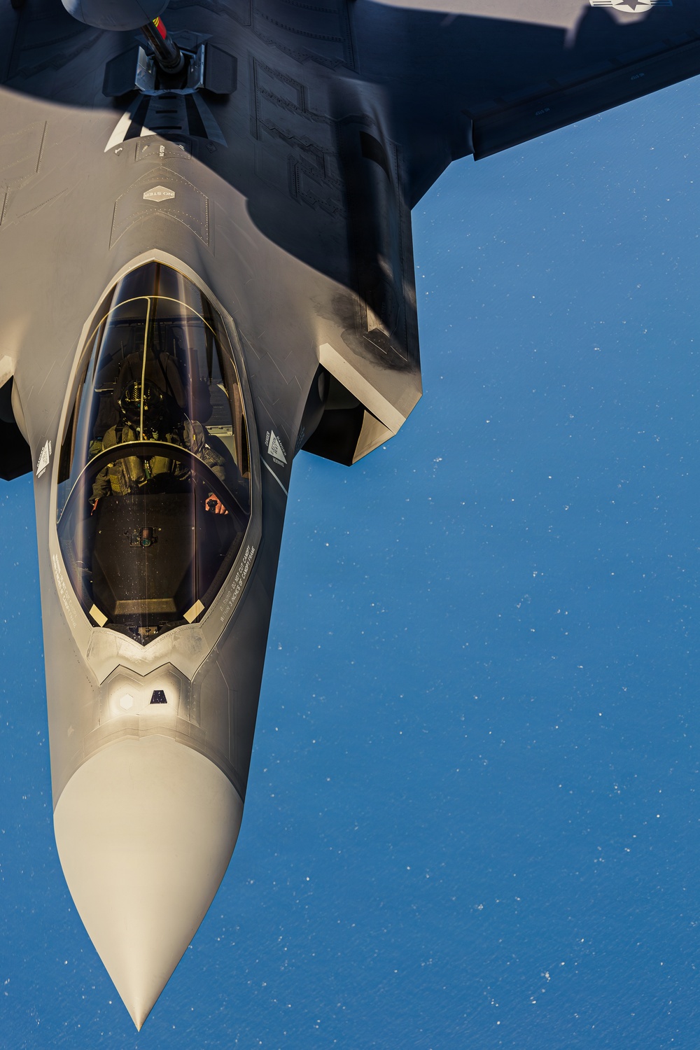 An aerial photograph showing a close-up view of an F-35 Lightning II stealth fighter jet during an aerial refueling operation. A pilot is visible inside the single-seat cockpit, and the refueling boom from a tanker aircraft is visible at the top of the frame. The image is taken from the perspective of the tanker aircraft looking down at the receiver jet against a clear blue sky.