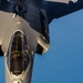 An aerial photograph showing a close-up view of an F-35 Lightning II stealth fighter jet during an aerial refueling operation. A pilot is visible inside the single-seat cockpit, and the refueling boom from a tanker aircraft is visible at the top of the frame. The image is taken from the perspective of the tanker aircraft looking down at the receiver jet against a clear blue sky.