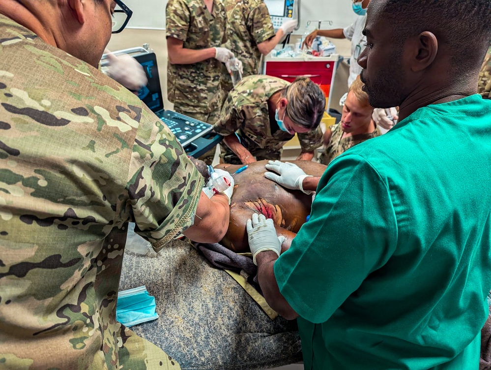 Military medical personnel and civilian healthcare workers are performing emergency trauma care on a patient in what appears to be a field or forward surgical setting. Multiple providers in camouflage uniforms and scrubs are working simultaneously on the patient, who has visible wounds. Medical equipment including a portable ultrasound device is visible in the background.