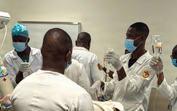 A medical team is gathered around a patient on a gurney in what appears to be a hospital or clinical setting. The group includes a person in military camouflage uniform alongside several medical personnel wearing white lab coats, surgical masks, and head coverings. The scene depicts active patient care or a medical training exercise involving IV administration and airway management.