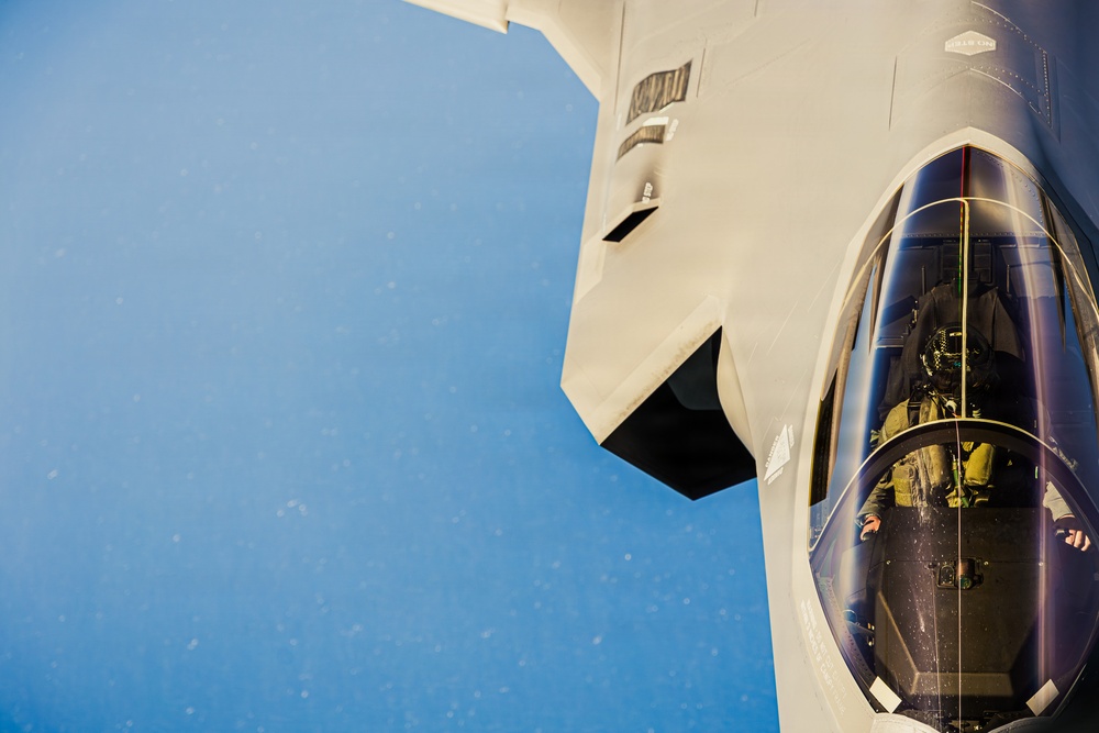 A close-up aerial photograph of a fifth-generation stealth fighter jet, identifiable as an F-35 Lightning II, captured from above and slightly to the side during flight. A pilot in full flight gear including helmet and oxygen mask is visible inside the cockpit. The image appears to have been taken from a tanker aircraft during or near an aerial refueling operation.