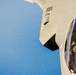 A close-up aerial photograph of a fifth-generation stealth fighter jet, identifiable as an F-35 Lightning II, captured from above and slightly to the side during flight. A pilot in full flight gear including helmet and oxygen mask is visible inside the cockpit. The image appears to have been taken from a tanker aircraft during or near an aerial refueling operation.