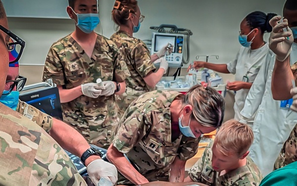 A medical team composed of military personnel in camouflage uniforms and civilian medical staff in white coats are performing emergency trauma care on a patient. Multiple providers are simultaneously working on the patient, suggesting a high-acuity resuscitation or trauma scenario. The setting appears to be a clinical treatment room equipped with medical monitoring equipment.
