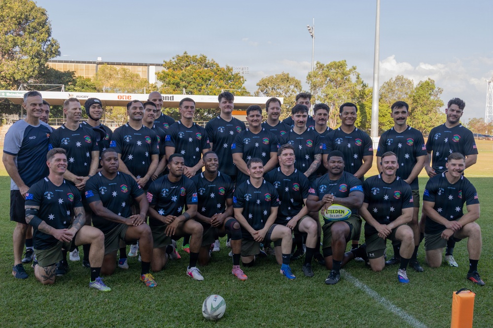 A rugby team poses for a group photo on a grass field at what appears to be a rugby stadium. The players are wearing matching dark navy jerseys featuring star emblems and both Australian and American flag patches on the sleeves, suggesting a joint or combined military/representative team. One player in the front row holds a Steeden rugby league ball.