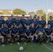 A rugby team poses for a group photo on a grass field at what appears to be a rugby stadium. The players are wearing matching dark navy jerseys featuring star emblems and both Australian and American flag patches on the sleeves, suggesting a joint or combined military/representative team. One player in the front row holds a Steeden rugby league ball.