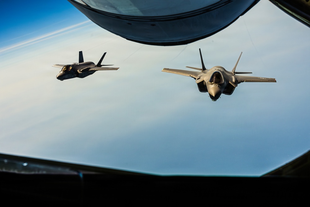Two F-35 Lightning II stealth fighter jets are photographed in formation flight from the boom operator's window of a tanker aircraft, likely a KC-135 or KC-46. The image is taken from below the tanker's fuselage looking outward, with the refueling boom visible at the top of the frame. The aircraft are flying at high altitude over a cloud layer with a visible horizon curvature.