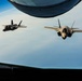 Two F-35 Lightning II stealth fighter jets are photographed in formation flight from the boom operator's window of a tanker aircraft, likely a KC-135 or KC-46. The image is taken from below the tanker's fuselage looking outward, with the refueling boom visible at the top of the frame. The aircraft are flying at high altitude over a cloud layer with a visible horizon curvature.