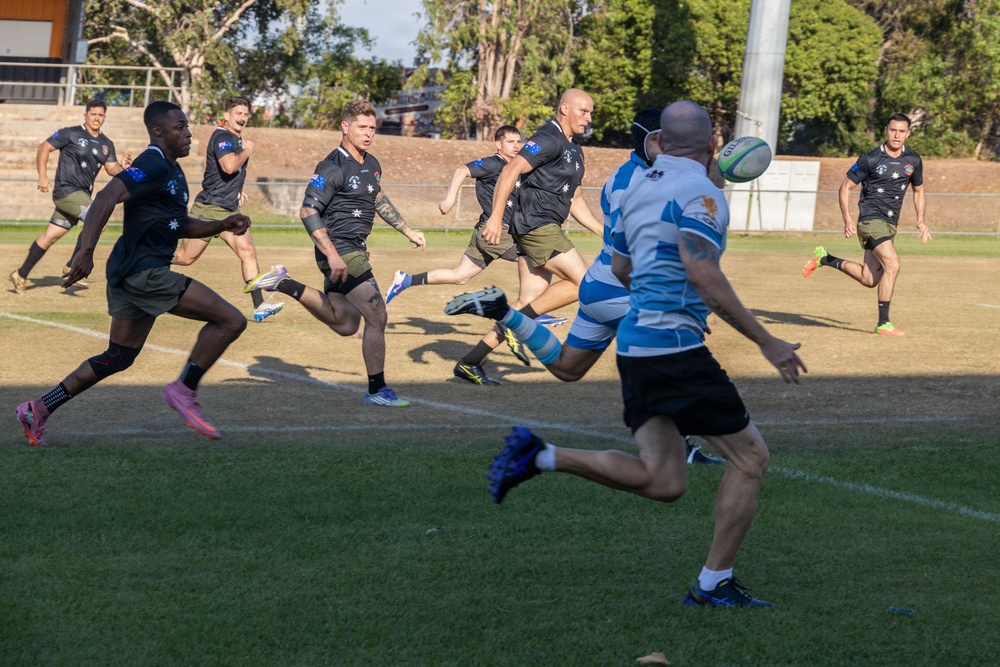 A rugby match is in progress on a grass field, with two teams competing — one wearing black uniforms with Australian flag patches and olive/khaki shorts, and the other in blue and white hooped jerseys. A player in the blue and white kit has just kicked the ball, which is visible in the air to the right. The action is dynamic with multiple players in full sprint across the well-maintained pitch.