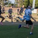 A rugby match is in progress on a grass field, with two teams competing — one wearing black uniforms with Australian flag patches and olive/khaki shorts, and the other in blue and white hooped jerseys. A player in the blue and white kit has just kicked the ball, which is visible in the air to the right. The action is dynamic with multiple players in full sprint across the well-maintained pitch.