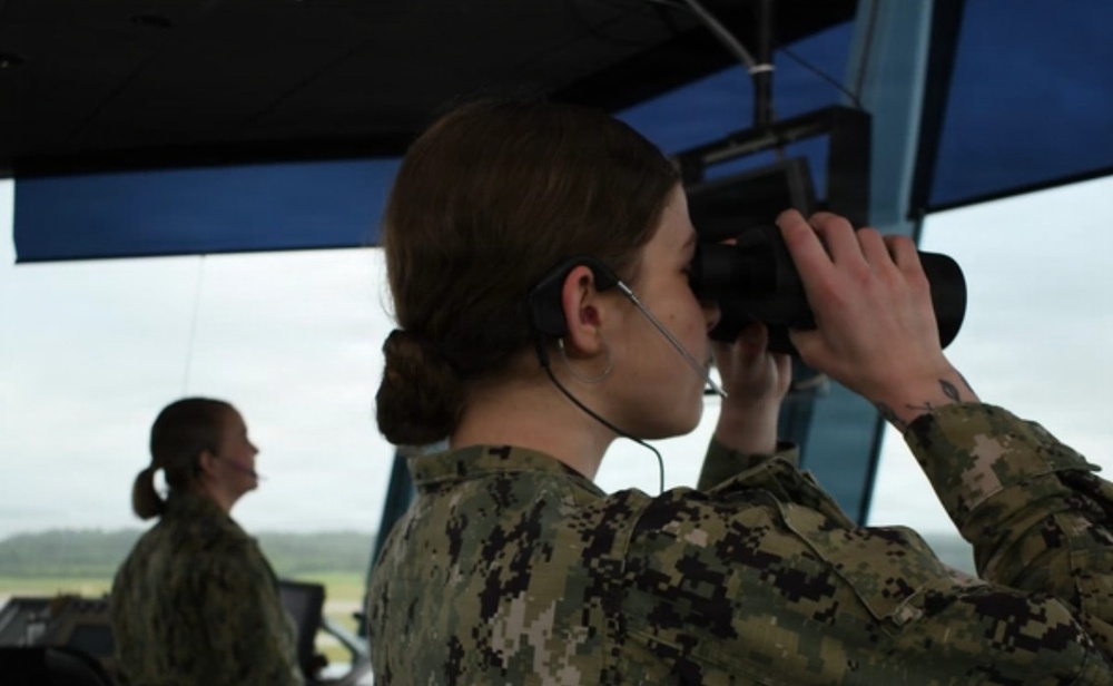 Two female military personnel are stationed in what appears to be an air traffic control tower. The foreground figure is using binoculars while wearing a communication headset, suggesting active observation duties. Both individuals are dressed in U.S. Navy Working Uniform (NWU) digital camouflage pattern.
