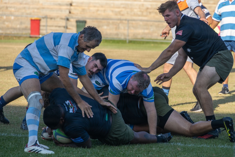 A rugby match is in progress with players from two teams engaged in a ruck or tackle situation on a grass field. One team wears blue and white hooped jerseys while the opposing team wears dark (black/olive) uniforms. Several players are piled on the ground competing for the ball while others stand over the ruck.