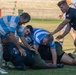 A rugby match is in progress with players from two teams engaged in a ruck or tackle situation on a grass field. One team wears blue and white hooped jerseys while the opposing team wears dark (black/olive) uniforms. Several players are piled on the ground competing for the ball while others stand over the ruck.