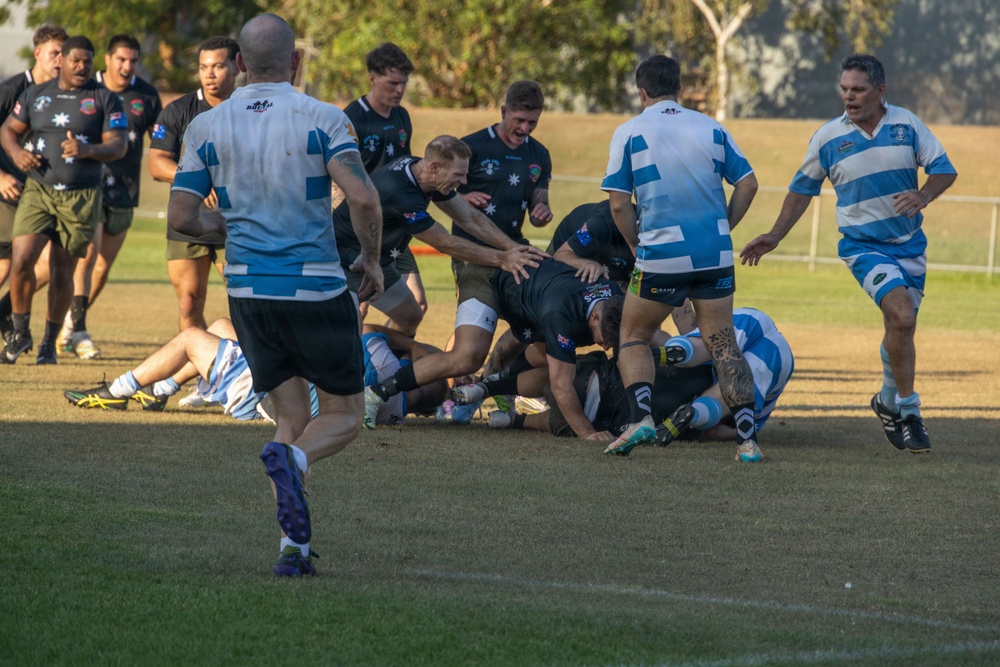 A rugby league or rugby union match is in progress on a grass field, featuring two teams — one in black uniforms with Australian flag patches and one in blue-and-white hooped jerseys. Players are engaged in a ruck or tackle situation near what appears to be the try line, with multiple players piling on. The image is taken during what appears to be late afternoon based on the warm, golden lighting.