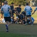 A rugby league or rugby union match is in progress on a grass field, featuring two teams — one in black uniforms with Australian flag patches and one in blue-and-white hooped jerseys. Players are engaged in a ruck or tackle situation near what appears to be the try line, with multiple players piling on. The image is taken during what appears to be late afternoon based on the warm, golden lighting.
