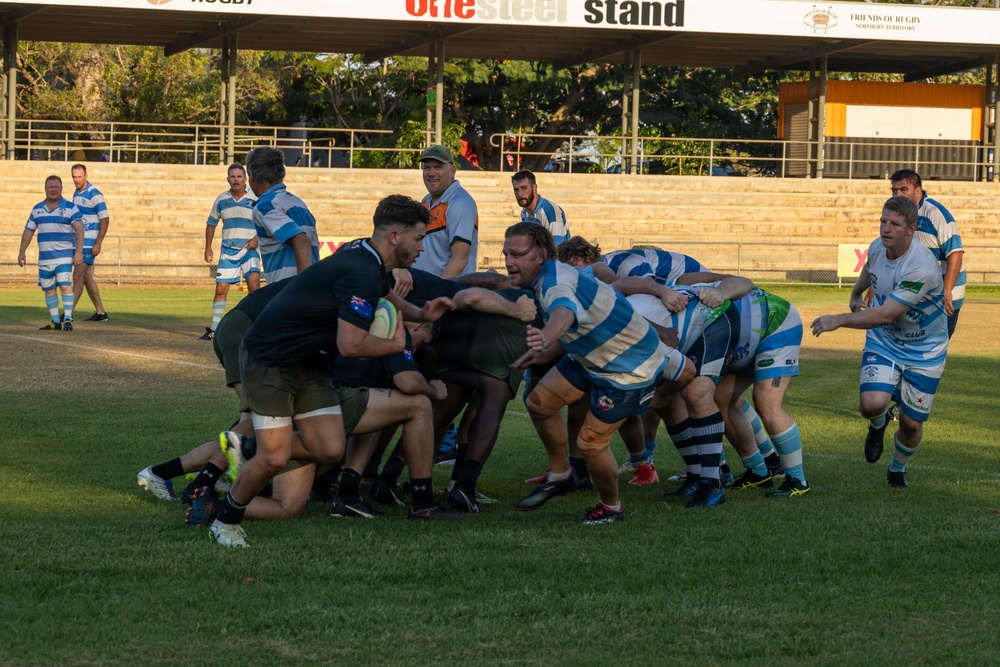 A rugby union match is being played at a ground in the Northern Territory, Australia, as indicated by the 'Friends of Rugby Northern Territory' signage. Two teams are engaged in a ruck or maul contest near the center of the field, with one team wearing black and the other in blue-and-white hooped jerseys. The match is taking place at an established club ground featuring a covered grandstand labeled the 'OneSteel Stand'.