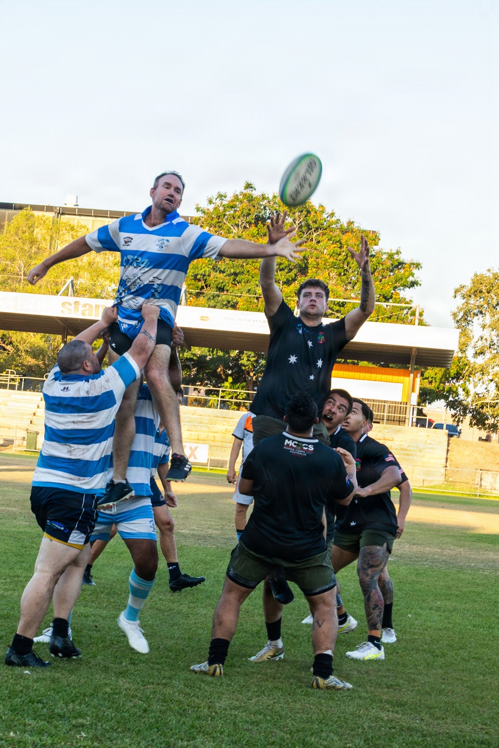 A rugby union lineout is being contested between two teams at an outdoor ground. A player in a blue and white hooped jersey is being lifted by teammates to contest the ball against opponents in black jerseys. The action is captured mid-air with the ball visible above the players.