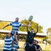 A rugby union lineout is being contested between two teams at an outdoor ground. A player in a blue and white hooped jersey is being lifted by teammates to contest the ball against opponents in black jerseys. The action is captured mid-air with the ball visible above the players.