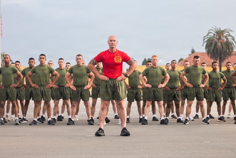 A military instructor or drill sergeant wearing a red t-shirt stands at the front of a large formation of recruits or trainees dressed in olive green physical training uniforms. All individuals have their hands on their hips in a uniform rest position, suggesting a physical training session or formation drill. The setting appears to be an outdoor parade ground or PT area, likely at a U.S. Marine Corps base based on the uniform style and insignia visible on the red shirt.