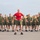 A military instructor or drill sergeant wearing a red t-shirt stands at the front of a large formation of recruits or trainees dressed in olive green physical training uniforms. All individuals have their hands on their hips in a uniform rest position, suggesting a physical training session or formation drill. The setting appears to be an outdoor parade ground or PT area, likely at a U.S. Marine Corps base based on the uniform style and insignia visible on the red shirt.