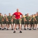 A military instructor or drill sergeant wearing a red t-shirt stands at the front of a large formation of recruits or trainees dressed in olive green physical training uniforms. All individuals have their hands on their hips in a uniform rest position, suggesting a physical training session or formation drill. The setting appears to be an outdoor parade ground or PT area, likely at a U.S. Marine Corps base based on the uniform style and insignia visible on the red shirt.