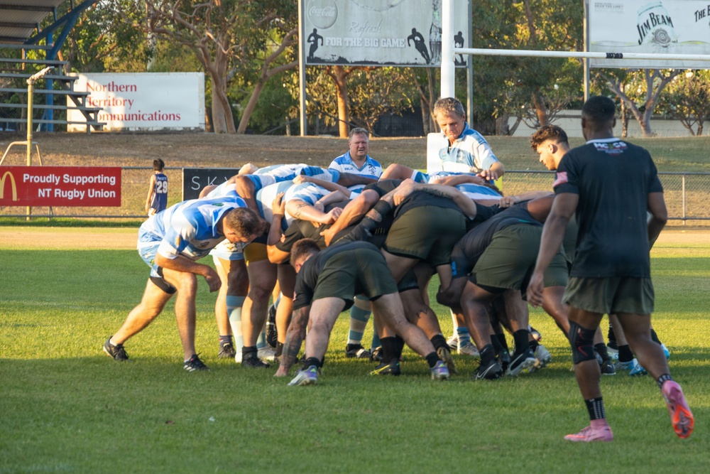 A rugby union scrum is taking place on a grass field during what appears to be a training session or match in the Northern Territory, Australia. Two teams are engaged — one in blue and white hooped jerseys and one in dark/black kit. Coaching staff and additional players observe from the sidelines near the goal posts.