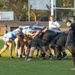 A rugby union scrum is taking place on a grass field during what appears to be a training session or match in the Northern Territory, Australia. Two teams are engaged — one in blue and white hooped jerseys and one in dark/black kit. Coaching staff and additional players observe from the sidelines near the goal posts.
