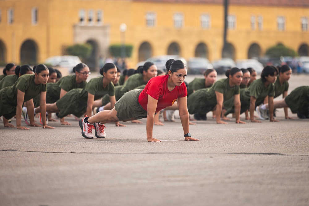 A group of female military recruits or trainees are performing push-ups in formation on a paved parade ground or drill area. A female instructor or drill sergeant wearing a red t-shirt leads the exercise from the front, distinguishing herself from the recruits in olive/green uniforms. A large yellow colonial-style building is visible in the background, consistent with a military base or academy.