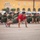 A group of female military recruits or trainees are performing push-ups in formation on a paved parade ground or drill area. A female instructor or drill sergeant wearing a red t-shirt leads the exercise from the front, distinguishing herself from the recruits in olive/green uniforms. A large yellow colonial-style building is visible in the background, consistent with a military base or academy.