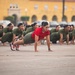 A group of female military recruits or trainees are performing push-ups in formation on a paved parade ground or drill area. A female instructor or drill sergeant wearing a red t-shirt leads the exercise from the front, distinguishing herself from the recruits in olive/green uniforms. A large yellow colonial-style building is visible in the background, consistent with a military base or academy.