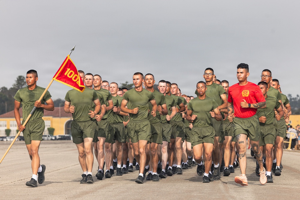 A large formation of military recruits in olive green PT (physical training) uniforms are running in formation on a paved parade deck or tarmac. A guidon bearer on the left carries a red and yellow flag with the number '100' visible. One individual in a red shirt, likely an instructor or drill instructor, runs alongside the formation.