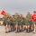 A large formation of military recruits in olive green PT (physical training) uniforms are running in formation on a paved parade deck or tarmac. A guidon bearer on the left carries a red and yellow flag with the number '100' visible. One individual in a red shirt, likely an instructor or drill instructor, runs alongside the formation.