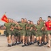 A large formation of military recruits in olive green PT (physical training) uniforms are running in formation on a paved parade deck or tarmac. A guidon bearer on the left carries a red and yellow flag with the number '100' visible. One individual in a red shirt, likely an instructor or drill instructor, runs alongside the formation.
