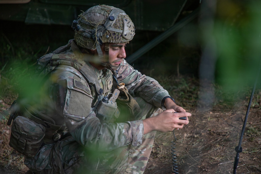 A U.S. Army soldier in full combat gear is crouched near a military vehicle in a wooded or field environment. The soldier appears to be operating a handheld radio or communication device, with a coiled antenna cable visible in the foreground. The image is captured through foliage, suggesting a field exercise or operational setting.