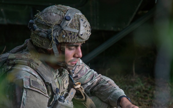 A U.S. Army soldier in full combat gear is crouched near a military vehicle in a wooded or field environment. The soldier appears to be operating a handheld radio or communication device, with a coiled antenna cable visible in the foreground. The image is captured through foliage, suggesting a field exercise or operational setting.
