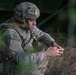 A U.S. Army soldier in full combat gear is crouched near a military vehicle in a wooded or field environment. The soldier appears to be operating a handheld radio or communication device, with a coiled antenna cable visible in the foreground. The image is captured through foliage, suggesting a field exercise or operational setting.