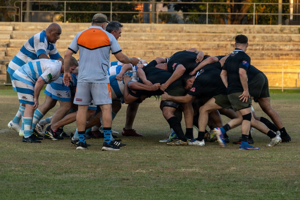 A rugby match is in progress featuring two teams engaged in a scrum or maul formation. One team wears blue and white hooped jerseys while the opposing team is dressed entirely in black. A referee in an orange and white vest is visible overseeing the play near the pack.