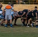 A rugby match is in progress featuring two teams engaged in a scrum or maul formation. One team wears blue and white hooped jerseys while the opposing team is dressed entirely in black. A referee in an orange and white vest is visible overseeing the play near the pack.